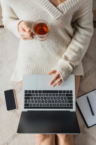 Вид зверху обрізаної жінки, що тримає скляну чашку з чаєм, використовуючи ноутбук біля смартфона з порожнім екраном — стокове фото
