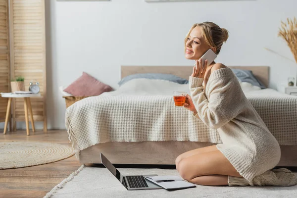 Весела жінка тримає скляну чашку з чаєм і розмовляє на смартфоні біля ноутбука на килимі — стокове фото