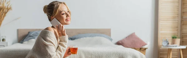 Веселая женщина, держа стеклянную чашку с чаем и разговаривая на смартфоне в спальне, баннер — стоковое фото