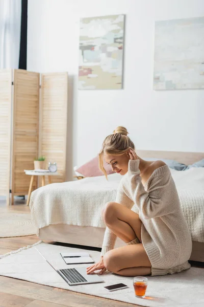 Очаровательная женщина в свитере с помощью ноутбука возле мобильного телефона и чашку чая на ковре в спальне — стоковое фото