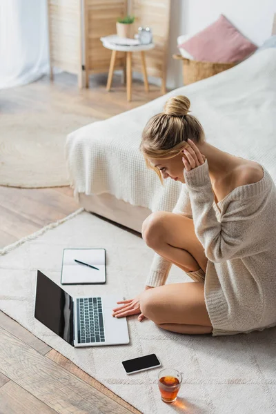 Mulher loira em suéter usando laptop perto de telefone celular e xícara de chá no tapete no quarto — Fotografia de Stock