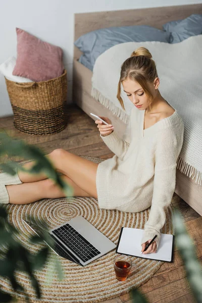 Hübsche Frau im Pullover, die neben Laptop Notizen macht und Smartphone hält, während sie neben dem Bett sitzt — Stockfoto
