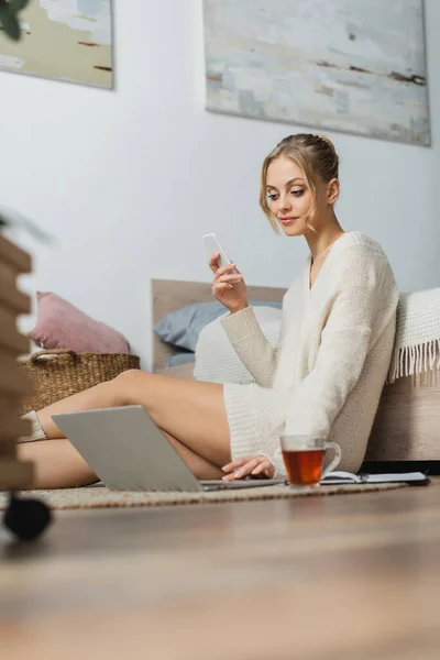 Donna bionda in maglione usando il computer portatile mentre tiene lo smartphone e sta seduta vicino al letto — Foto stock