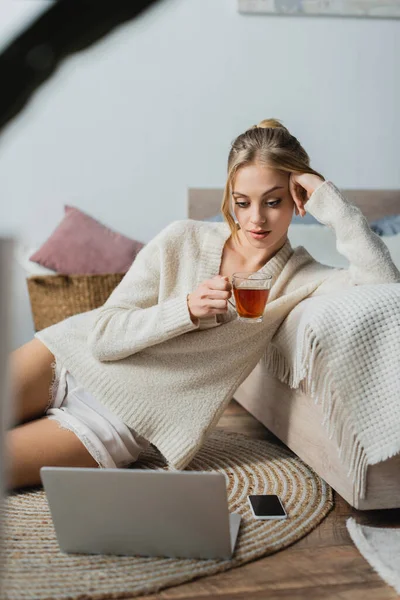 Young blonde woman in sweater holding glass cup with tea and looking at laptop in bedroom — Stock Photo