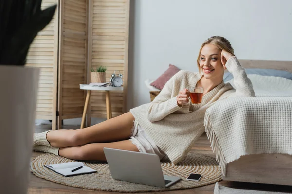 Усміхнена блондинка в светрі тримає скляну чашку з чаєм, сидячи біля гаджетів у спальні — стокове фото