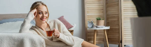 Sorrindo mulher loira em suéter segurando copo de vidro com chá no quarto, banner — Fotografia de Stock