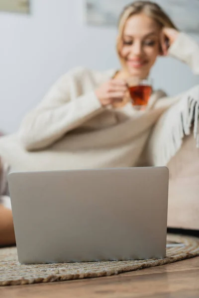 Mulher desfocada em suéter segurando copo de vidro com chá e olhando para laptop — Fotografia de Stock
