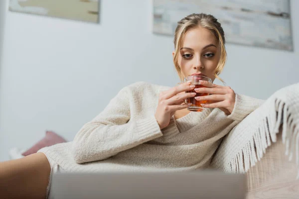 Blonde woman in sweater holding glass cup with tea and looking at blurred laptop in bedroom — Stock Photo