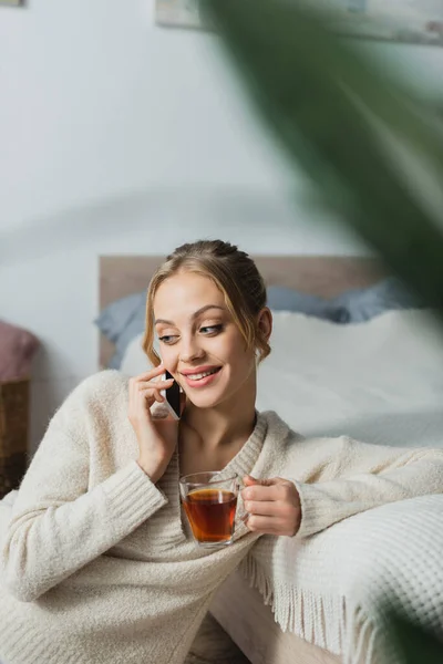 Mujer satisfecha hablando en el teléfono inteligente y sosteniendo una taza de té en el dormitorio - foto de stock