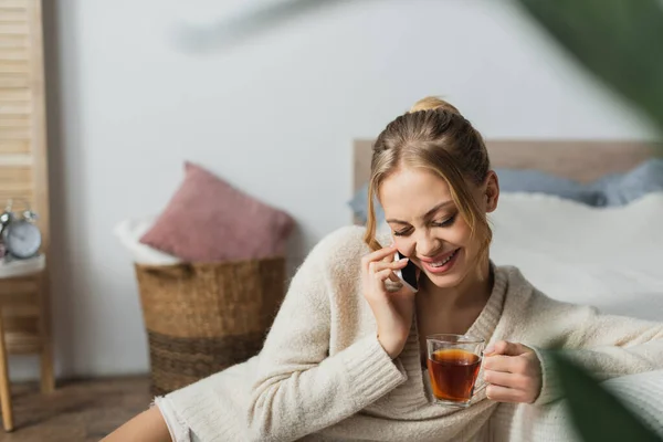 Mujer complacida hablando en el teléfono inteligente y sosteniendo una taza de té en el dormitorio - foto de stock