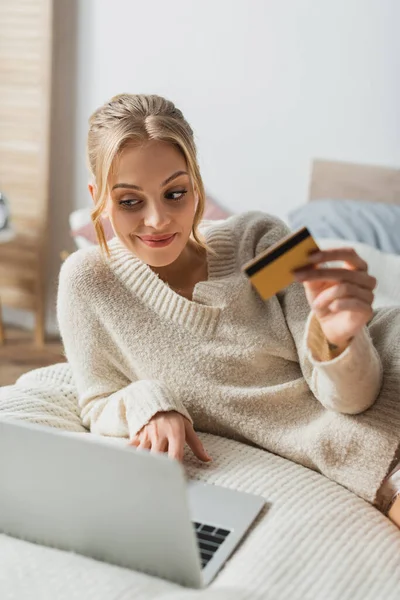 Mujer alegre que sostiene la tarjeta de crédito y el uso de la computadora portátil mientras que hace compras en línea en dormitorio - foto de stock