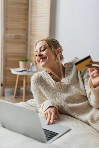 Задоволена жінка тримає кредитну картку і використовує ноутбук під час онлайн-покупки в спальні — стокове фото