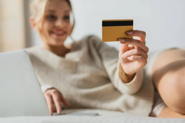 Размытая женщина, держащая кредитную карту и используя ноутбук во время онлайн-покупок в спальне — стоковое фото