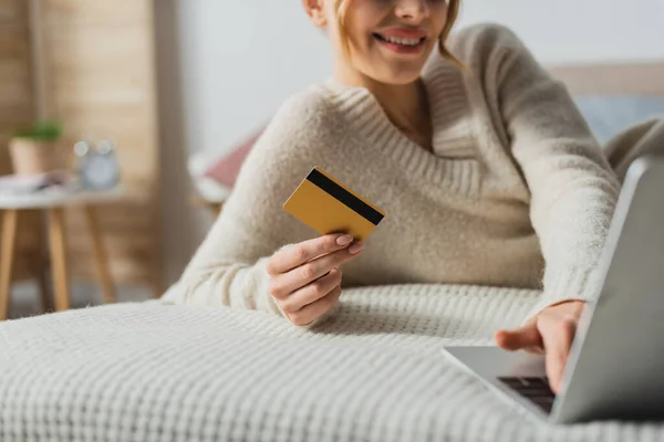 Частковий вид веселої жінки, що тримає кредитну картку і використовує ноутбук під час онлайн-покупки в спальні — стокове фото