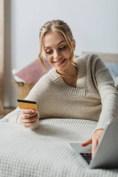 Zufriedene Frau im Pullover mit Kreditkarte und Laptop beim Online-Shopping im Schlafzimmer — Stockfoto