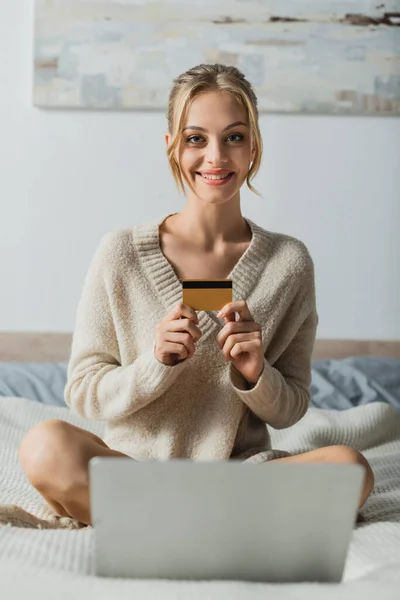Веселая женщина, держащая кредитную карту возле ноутбука во время покупок в спальне — стоковое фото