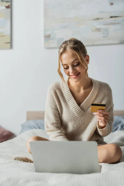 Mujer satisfecha sosteniendo la tarjeta de crédito cerca del ordenador portátil mientras que hace compras en línea en dormitorio - foto de stock