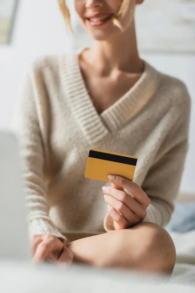 Abgeschnittene Ansicht einer lächelnden Frau, die ihre Kreditkarte in der Nähe ihres Laptops hält und auf dem Bett sitzt — Stockfoto