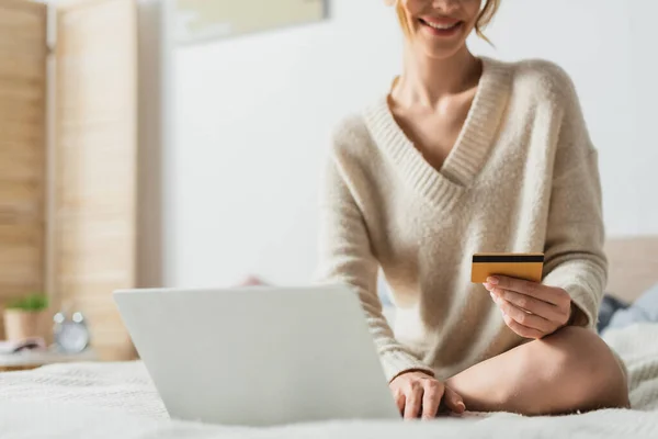Vue partielle de la femme heureuse tenant la carte de crédit près de l'ordinateur portable tout en faisant des achats en ligne dans la chambre — Photo de stock