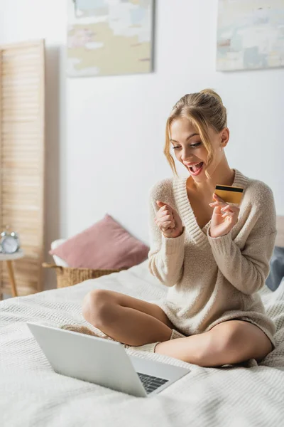 Erstaunte Frau mit Kreditkarte in der Nähe von Laptop beim Online-Shopping im Schlafzimmer — Stockfoto