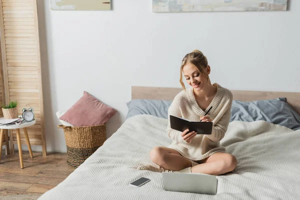 Cheerful blonde woman taking notes near gadgets on bed in modern apartment — Stock Photo