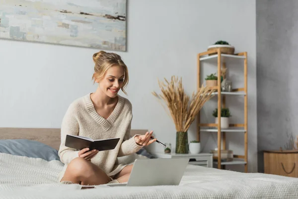 Cheerful young woman holding notebook and pen while watching webinar on laptop in bedroom — Stock Photo