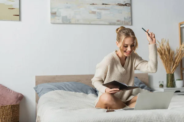 Веселая молодая женщина с ноутбуком и ручкой во время учебы в Интернете в спальне — стоковое фото