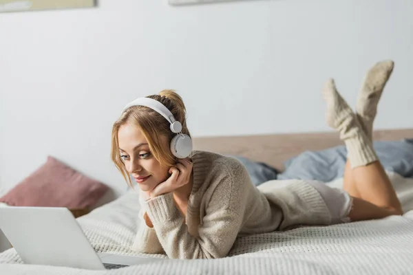Young blonde woman in wireless headphones watching movie on laptop in bedroom — Stock Photo