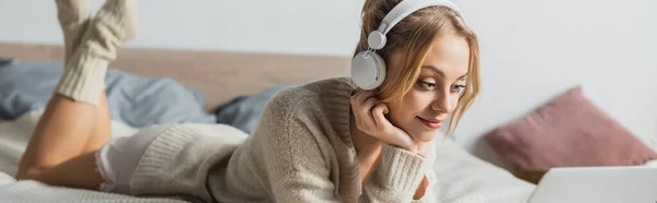 Mujer rubia joven en auriculares inalámbricos viendo la película en el ordenador portátil en el dormitorio, pancarta - foto de stock