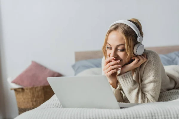Весела жінка в бездротових навушниках дивиться комедійний фільм на ноутбуці в спальні — стокове фото