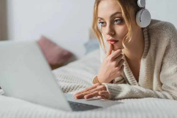 Зосереджена жінка в бездротових навушниках, використовуючи ноутбук у спальні — стокове фото