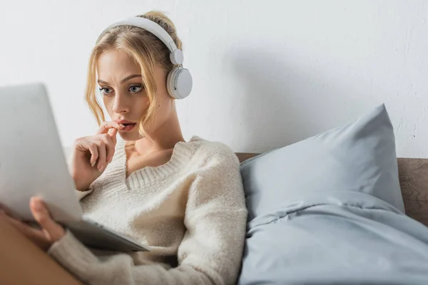 Зосереджена молода жінка в бездротових навушниках, використовуючи ноутбук у спальні — стокове фото