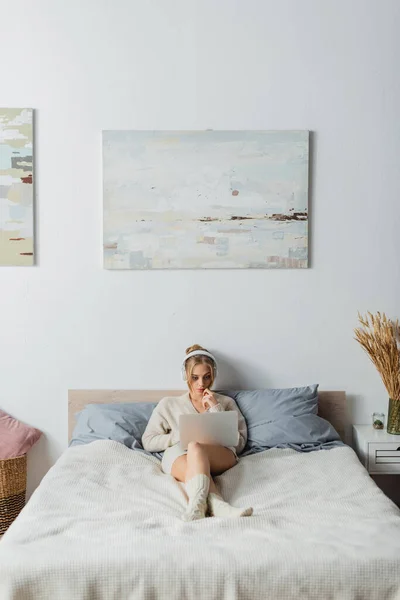Полная длина сфокусированной молодой женщины в беспроводных наушниках с помощью ноутбука в современной спальне — стоковое фото