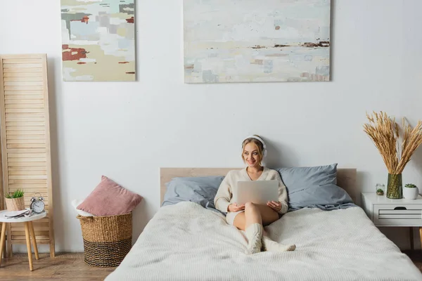 Volle Länge der fröhlichen jungen Frau in drahtlosen Kopfhörern mit Laptop im modernen Schlafzimmer — Stockfoto