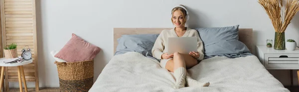 Piena lunghezza della giovane donna allegra in cuffie senza fili utilizzando il computer portatile in camera da letto moderna, banner — Foto stock