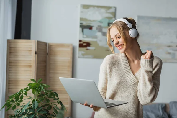 Freudige junge Frau mit drahtlosen Kopfhörern, die Musik hört und Laptop im modernen Schlafzimmer hält — Stockfoto