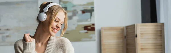 Радостная молодая женщина в беспроводных наушниках слушает музыку в современной квартире, баннер — стоковое фото