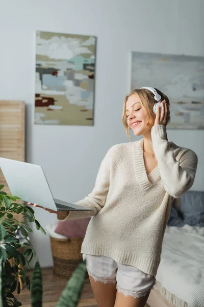 Mujer joven feliz en auriculares inalámbricos escuchando música y sosteniendo el ordenador portátil en el dormitorio moderno - foto de stock