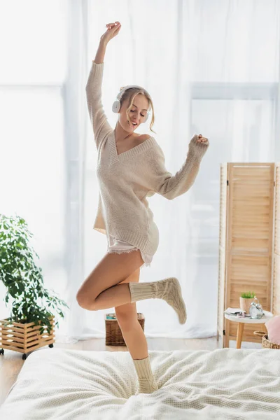 Полная длина радостной молодой женщины в беспроводных наушниках танцует на кровати и веселится в выходные дни — стоковое фото