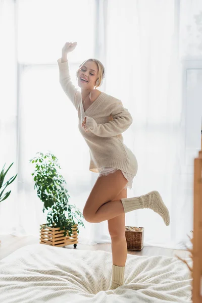 Piena lunghezza di donna spensierata in cuffie wireless ballare sul letto e divertirsi durante il fine settimana — Foto stock