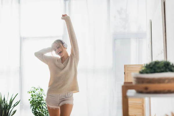 Задоволена жінка в в'язаному светрі і бездротових навушниках танцює, розважаючись в сучасній квартирі — стокове фото