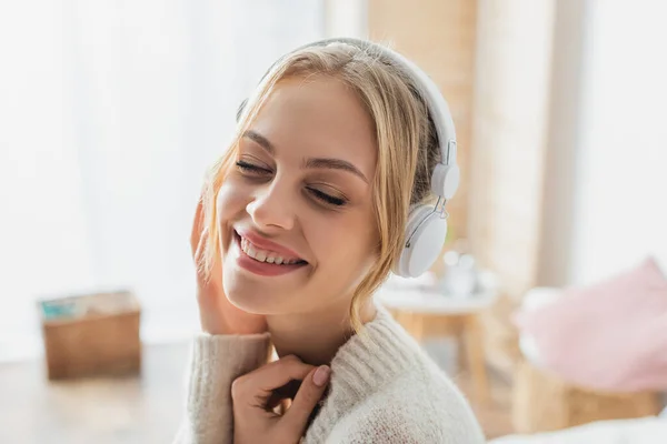 Mujer joven llena de alegría escuchando música en auriculares inalámbricos mientras sonríe con los ojos cerrados - foto de stock