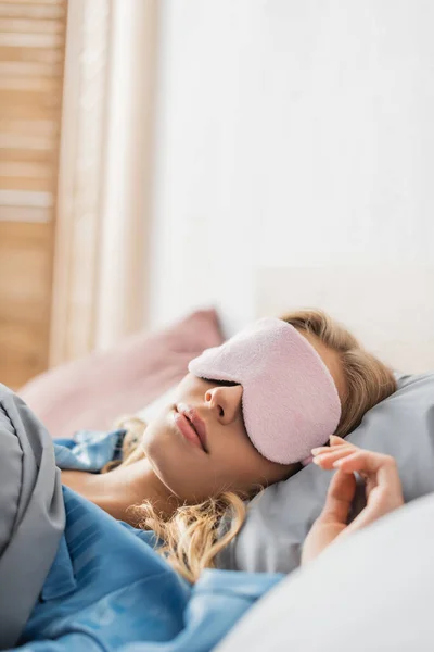 Femme blonde en masque de sommeil rose et pyjama bleu reposant au lit — Photo de stock