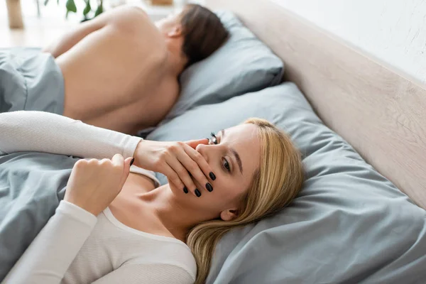 Blonde woman covering mouth while waking up with shirtless man in her bed after one night stand — Stock Photo