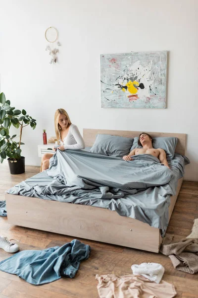 Blonde woman sitting on bed and pulling blanket while shirtless man sleeping after one night stand — Stock Photo