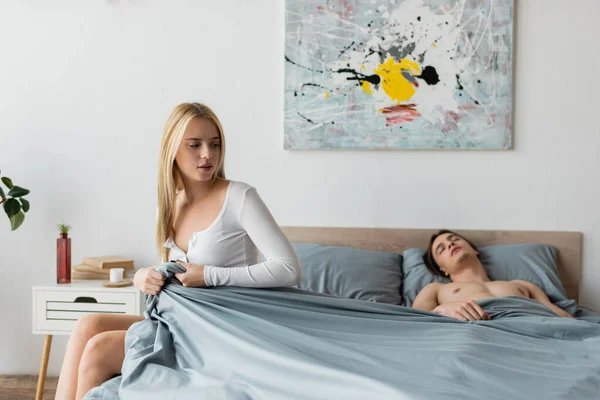 Blonde woman pulling blanket while shirtless man sleeping after one night stand — Stock Photo