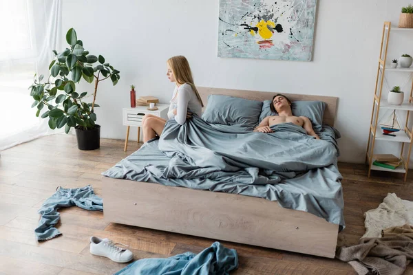 Розчарована жінка сидить на ліжку біля чоловіка після однієї ночі стоїть в безладному спальні — стокове фото