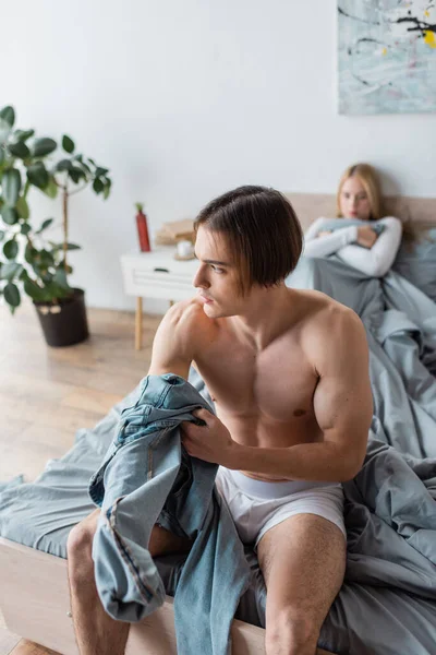 Без рубашки мужчина держит джинсы, сидя в постели рядом с женщиной после одной ночи — стоковое фото