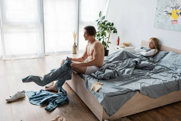 Full length of shirtless man in underwear wearing jeans near displeased woman in bed after one night stand — Stock Photo