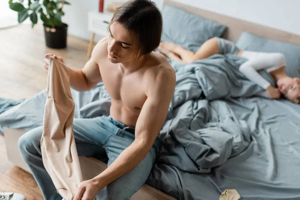 Без рубашки мужчина держит одежду, сидя на кровати рядом с женщиной после одной ночи — стоковое фото